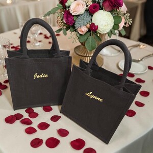 May include: Two black square tote bags made of a textured fabric, each featuring two sturdy handles. The bag on the left has the name Jodie embroidered in gold thread, and the bag on the right has the name Lynne embroidered in gold thread.