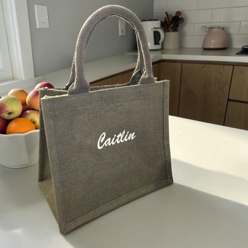 May include: A small, rectangular, olive-green burlap tote bag with two matching fabric handles. The front of the bag features the name 'Caitlin' embroidered in white cursive script lettering. The bag has a structured base and a textured weave.