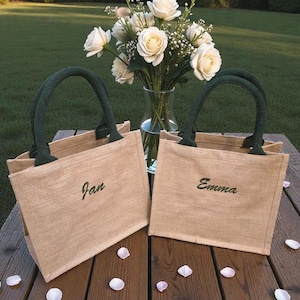 May include: Two rectangular tan jute tote bags with dark green handles. Each bag features a name embroidered in dark green cursive script on the front, with one reading Jan and the other reading Emma. The bags are positioned on a wooden surface.