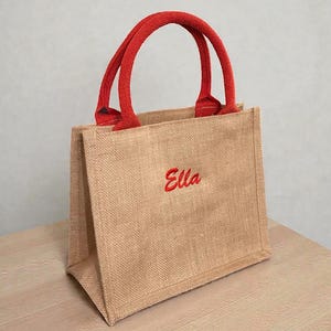 May include: A small tote bag made of tan jute fabric with two red woven handles. The front of the bag features the name Ella embroidered in red script lettering. The bag has a structured rectangular shape with a flat base and vertical side panels.
