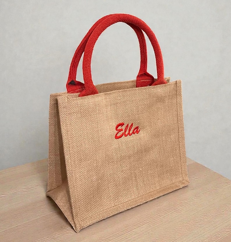 May include: A small tote bag made of tan jute fabric with two red woven handles. The front of the bag features the name Ella embroidered in red script lettering. The bag has a structured rectangular shape with a flat base and vertical side panels.