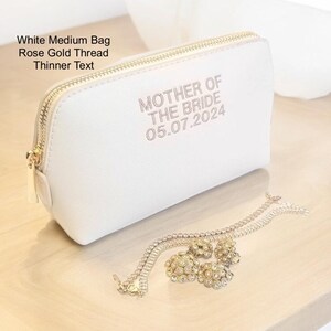 May include: A white medium bag with a rose gold zipper and text that reads "MOTHER OF THE BRIDE 05.07.2024". A gold-colored bracelet and earrings are next to the bag on a wooden surface.
