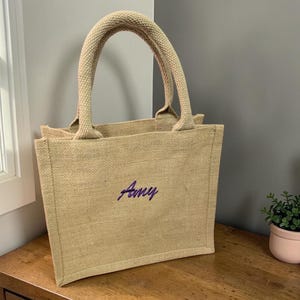 May include: A rectangular beige jute tote bag with two sturdy top handles. The front of the bag features the name Amy embroidered in a dark purple script font. The bag has a structured, flat-bottomed design with a visible woven texture.