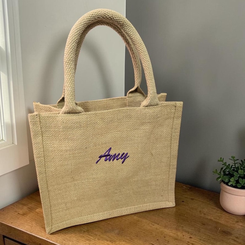 May include: A rectangular beige jute tote bag with two sturdy top handles. The front of the bag features the name Amy embroidered in a dark purple script font. The bag has a structured, flat-bottomed design with a visible woven texture.