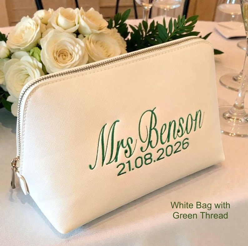 May include: A white cosmetic bag with a silver zipper and green embroidered text that reads "Mrs Benson 21.08.2026". The bag is rectangular and sits on a white surface, with a bouquet of white roses in the background.