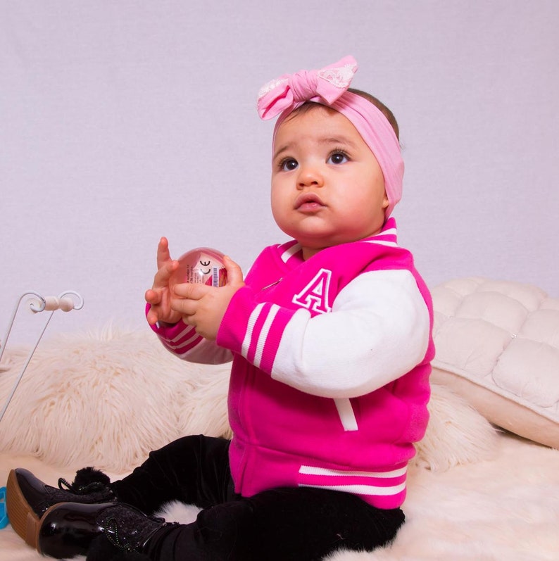 May include: A young child wearing a pink and white varsity jacket with a white letter 'A' on the sleeve. The child is holding a clear plastic ball and wearing a pink bow in their hair.