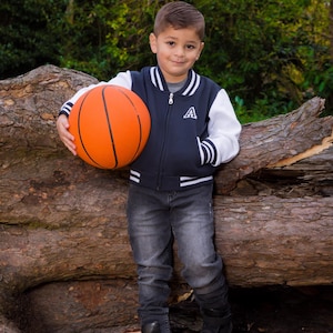 May include: A young boy wearing a navy blue and white baseball jacket with a white letter 'A' on the front, holds an orange basketball in front of a tree trunk.