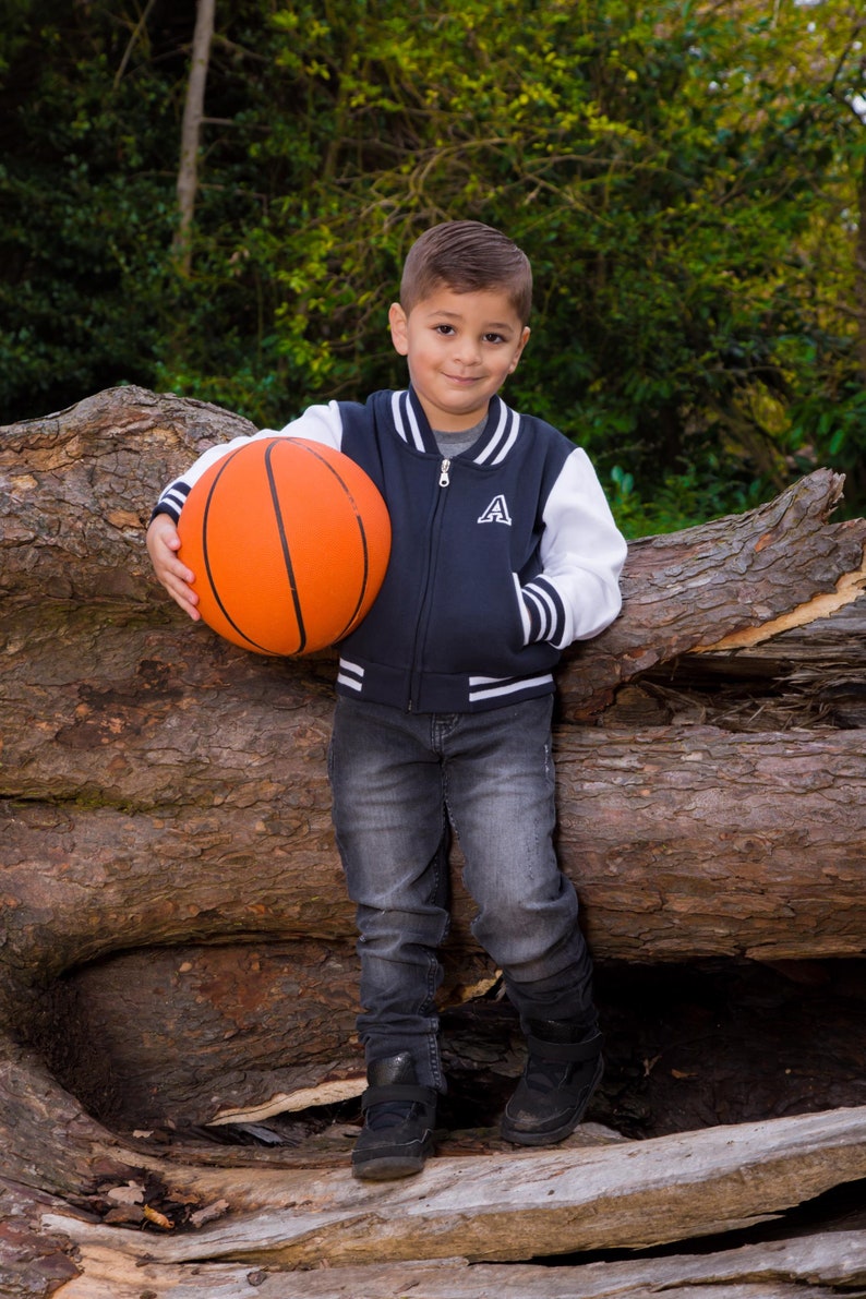 May include: A young boy wearing a navy blue and white baseball jacket with a white letter 'A' on the front, holds an orange basketball in front of a tree trunk.