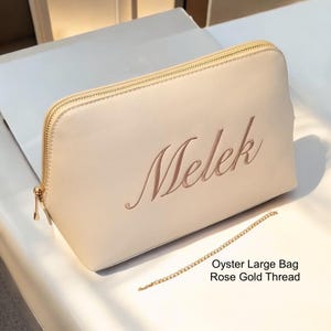 May include: A cream-colored cosmetic bag with a gold zipper and a rose gold embroidered name, "Melek." The bag is rectangular and has a matching gold chain. The text "Oyster Large Bag Rose Gold Thread" is also visible.