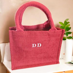 May include: A rectangular tote bag made of a textured, deep red woven fabric. The bag features two sturdy, rounded handles in the same red material and the white capital letters DD are centered on the front face of the bag in a serif font.