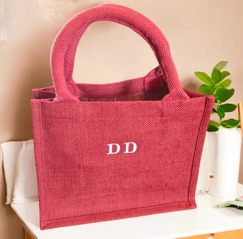 May include: A rectangular tote bag made of a textured, deep red woven fabric. The bag features two sturdy, rounded handles in the same red material and the white capital letters DD are centered on the front face of the bag in a serif font.