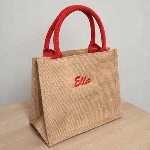 May include: A small tote bag made of tan jute fabric with two red woven handles. The front of the bag features the name Ella embroidered in red script lettering. The bag has a structured rectangular shape with a flat base and vertical side panels.