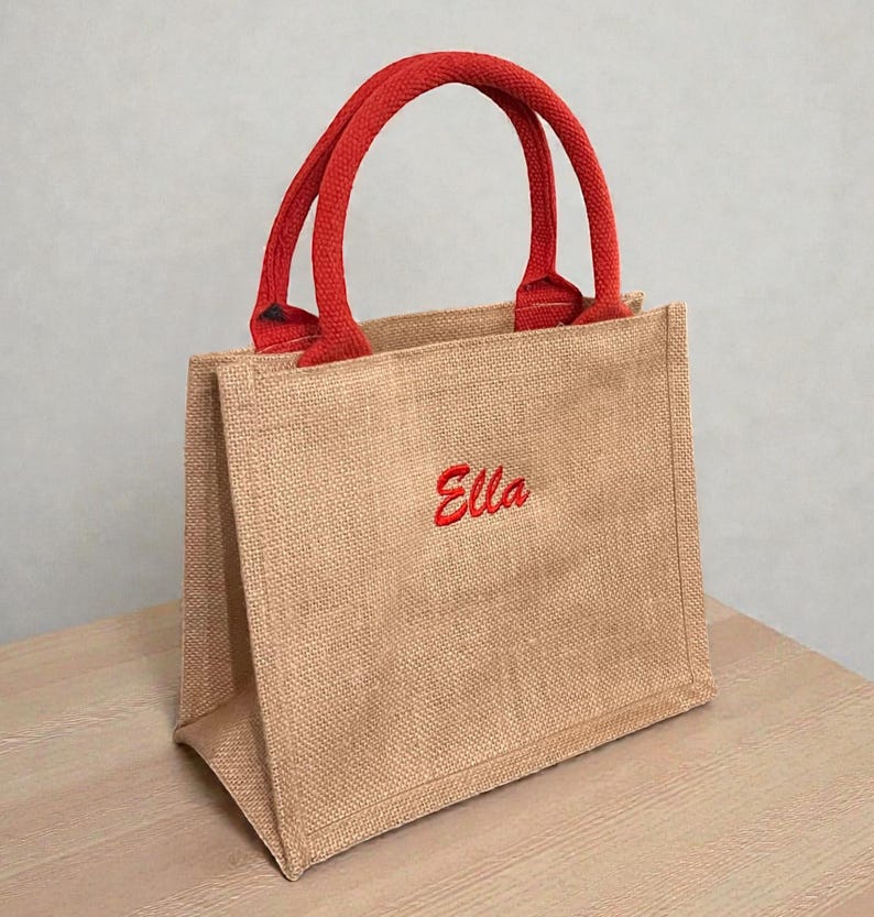 May include: A small tote bag made of tan jute fabric with two red woven handles. The front of the bag features the name Ella embroidered in red script lettering. The bag has a structured rectangular shape with a flat base and vertical side panels.