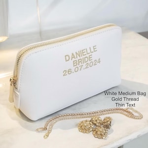 May include: A white medium bag with a gold zipper and gold text that reads "DANIELLE BRIDE 26.07.2024". A gold necklace and earrings are in front of the bag. The bag is on a white surface.