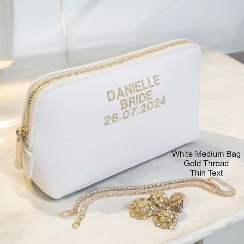 May include: A white medium bag with a gold zipper and gold text that reads "DANIELLE BRIDE 26.07.2024". A gold necklace and earrings are in front of the bag. The bag is on a white surface.