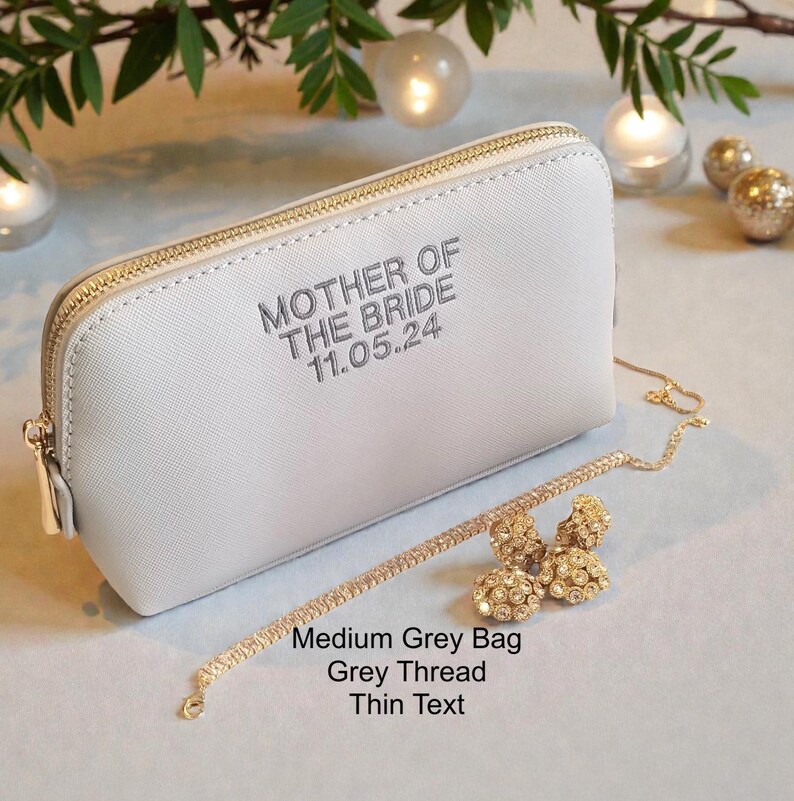 May include: A medium grey zippered bag with the text "MOTHER OF THE BRIDE 11.05.24" in grey thread. A gold-colored bracelet and earrings are next to the bag. The bag has a gold zipper.