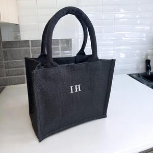 May include: A black tote bag made of a textured woven material featuring two sturdy handles and the letters IH embroidered in white on the front center. The bag has a structured rectangular shape with a flat base and vertical side panels.