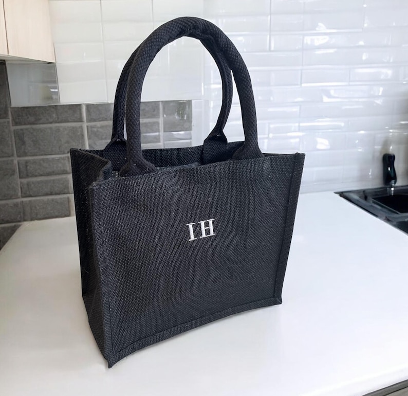 May include: A black tote bag made of a textured woven material featuring two sturdy handles and the letters IH embroidered in white on the front center. The bag has a structured rectangular shape with a flat base and vertical side panels.