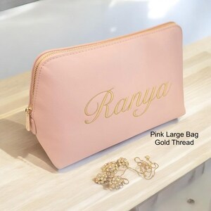 May include: A light pink cosmetic bag with a gold zipper and the name "Ranya" embroidered in gold thread. The bag is rectangular and sits on a wooden surface. A gold necklace and earrings are next to the bag.