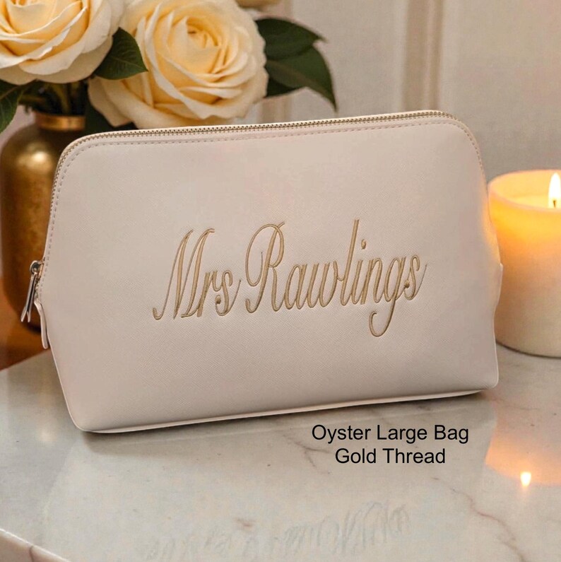 May include: An oyster-colored cosmetic bag with a gold zipper and the embroidered name "Mrs Rawlings" in gold thread. The bag is labeled "Oyster Large Bag Gold Thread". The bag is on a marble surface.