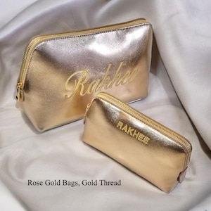 May include: Two rose gold cosmetic bags with gold zippers. The larger bag has the name "Rakhee" embroidered in gold thread. The smaller bag has the name "RAKHEE" embroidered in gold thread. The bags are on a satin surface.