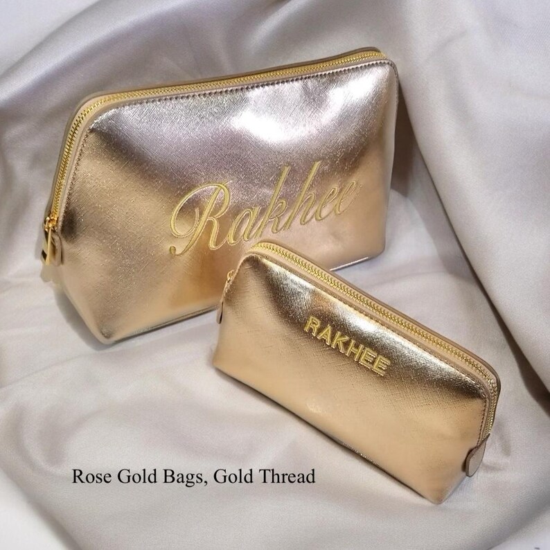 May include: Two rose gold cosmetic bags with gold zippers. The larger bag has the name "Rakhee" embroidered in gold thread. The smaller bag has the name "RAKHEE" embroidered in gold thread. The bags are on a satin surface.
