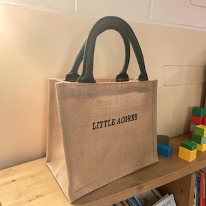 May include: A tan jute tote bag with two dark green fabric handles. The front of the bag features the text "LITTLE ACORNS" printed in dark green capital letters. The bag has a structured, rectangular shape with a visible woven texture.