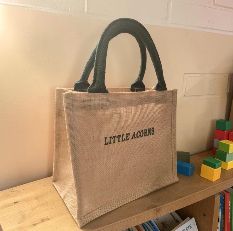 May include: A tan jute tote bag with two dark green fabric handles. The front of the bag features the text "LITTLE ACORNS" printed in dark green capital letters. The bag has a structured, rectangular shape with a visible woven texture.