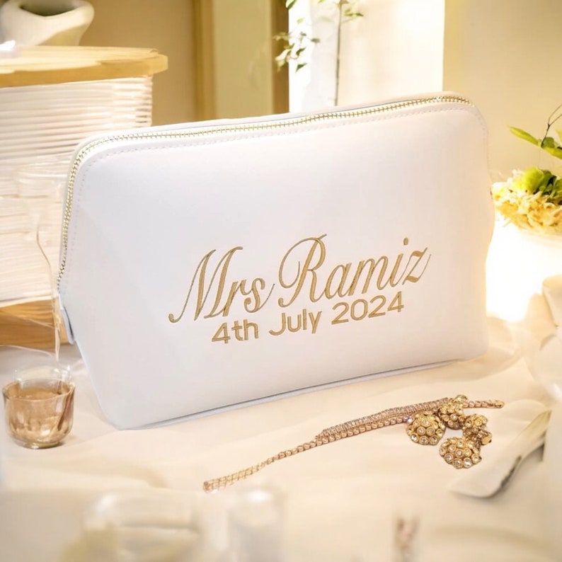May include: White cosmetic bag with a gold zipper and the inscription "Mrs Ramiz 4th July 2024" in gold script. A gold bracelet and earrings are next to the bag. The bag is on a white surface.