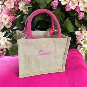 May include: A small, rectangular, natural-colored jute tote bag featuring two bright pink fabric handles and the name "Sanel" embroidered in matching pink thread on the front center of the bag, which has a structured, open-top design.