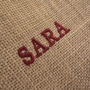 May include: A coarse, tan-colored burlap fabric featuring the name SARA embroidered in a dark red thread. The embroidery is positioned diagonally across the woven, textured surface of the material, showcasing the visible weave of the fibers.