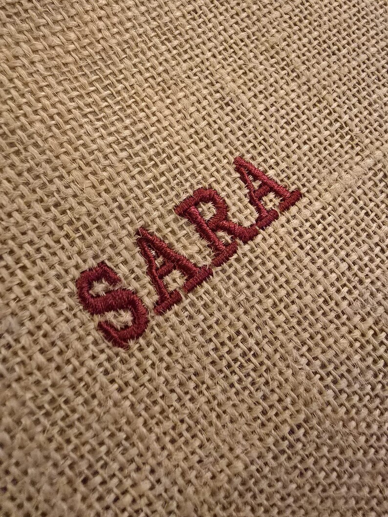 May include: A coarse, tan-colored burlap fabric featuring the name SARA embroidered in a dark red thread. The embroidery is positioned diagonally across the woven, textured surface of the material, showcasing the visible weave of the fibers.