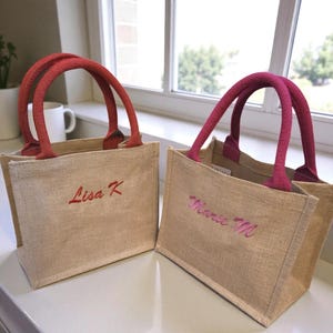 May include: Two small rectangular jute tote bags with open tops. The left bag has red handles and the text Lisa K embroidered in red. The right bag has magenta handles and the text Marie M embroidered in magenta. Both bags are made of tan fabric.