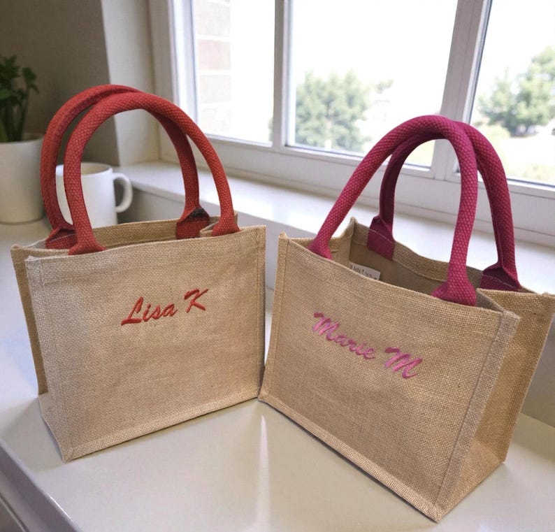 May include: Two small rectangular jute tote bags with open tops. The left bag has red handles and the text Lisa K embroidered in red. The right bag has magenta handles and the text Marie M embroidered in magenta. Both bags are made of tan fabric.