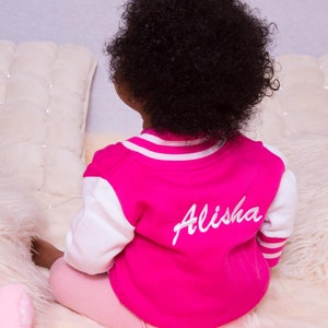 May include: A pink and white baseball jacket with the name "Alisha" embroidered on the back. The jacket is being worn by a doll with curly black hair.