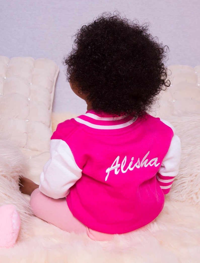 May include: A pink and white baseball jacket with the name "Alisha" embroidered on the back. The jacket is being worn by a doll with curly black hair.