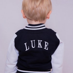 May include: A young child wearing a navy blue and white baseball jacket with the name "LUKE" embroidered on the back. The jacket has white sleeves and a white stripe around the collar and bottom.