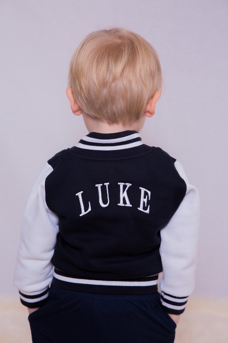 May include: A young child wearing a navy blue and white baseball jacket with the name "LUKE" embroidered on the back. The jacket has white sleeves and a white stripe around the collar and bottom.