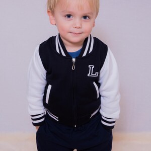 May include: A young boy wearing a navy blue and white baseball jacket with a white "L" on the front. He is wearing dark blue sweatpants and has blonde hair.