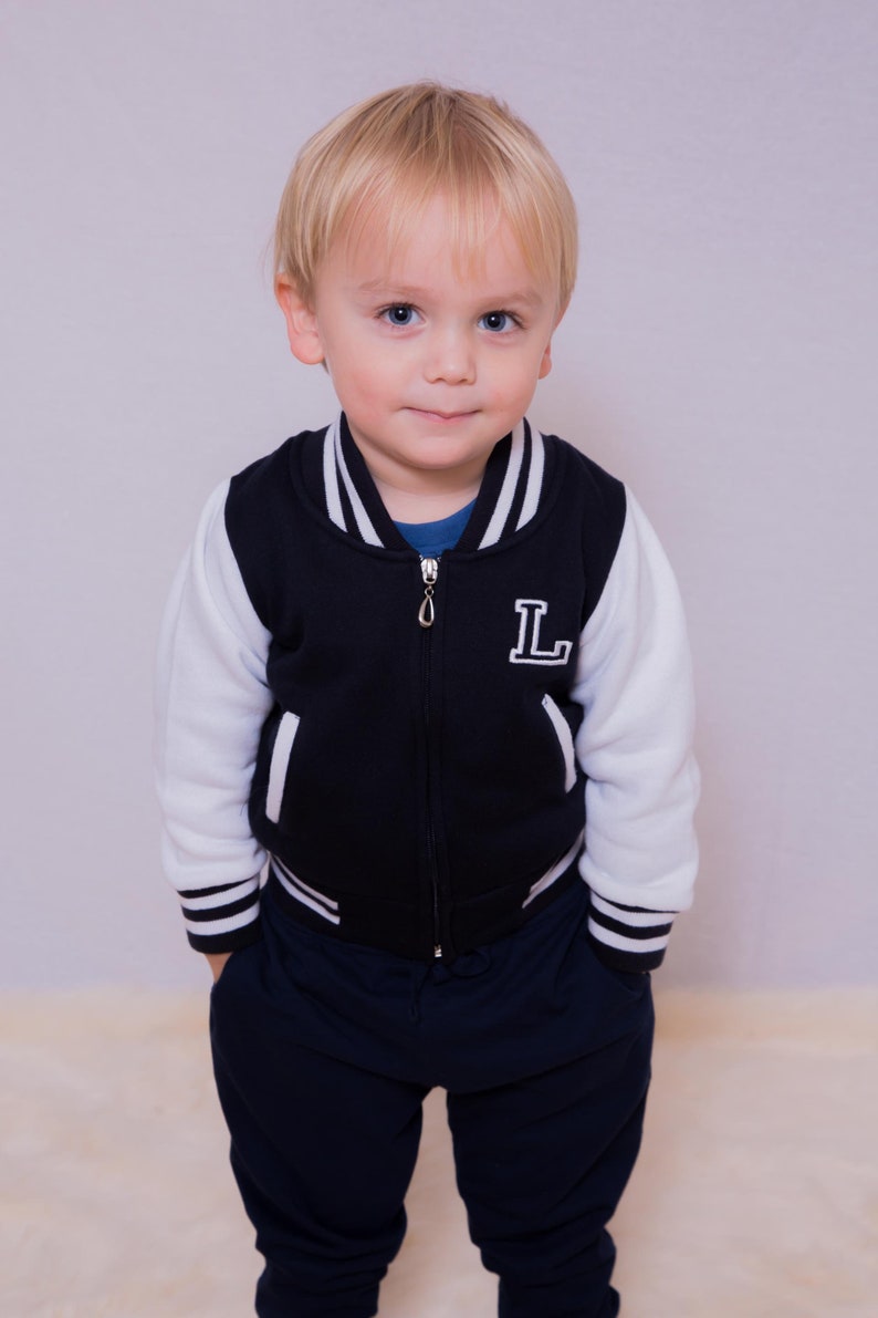 May include: A young boy wearing a navy blue and white baseball jacket with a white "L" on the front. He is wearing dark blue sweatpants and has blonde hair.
