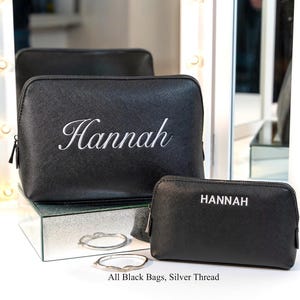 May include: Black cosmetic bags with silver thread embroidery. One bag has "Hannah" in cursive, the other has "HANNAH" in block letters. The bags are displayed with two silver bracelets. Text on the image reads: "All Black Bags, Silver Thread."