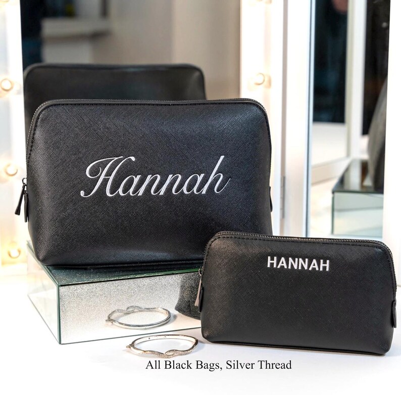 May include: Black cosmetic bags with silver thread embroidery. One bag has "Hannah" in cursive, the other has "HANNAH" in block letters. The bags are displayed with two silver bracelets. Text on the image reads: "All Black Bags, Silver Thread."