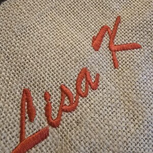 May include: A piece of light brown, coarse-woven burlap fabric featuring the name Lisa K embroidered in a vibrant, dark orange thread. The text is stitched in a cursive script font, standing out against the textured, natural fiber background.