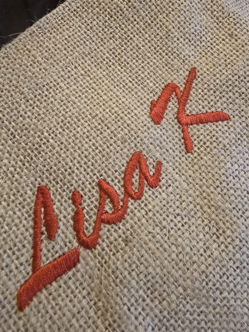 May include: A piece of light brown, coarse-woven burlap fabric featuring the name Lisa K embroidered in a vibrant, dark orange thread. The text is stitched in a cursive script font, standing out against the textured, natural fiber background.