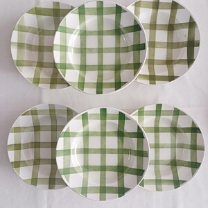 Könnte beinhalten: Sechs weiße Keramikplatten mit grünem Karomuster. Die Teller haben eine flache Schalenform und ein schlichtes Design. Das grüne Karomuster ist gleichmäßig verteilt und bedeckt die gesamte Oberfläche.