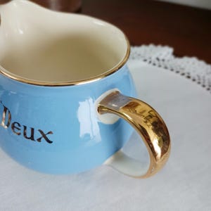 May include: A light blue creamer with a gold rim and handle. The word "Deux" is written in gold on the front of the creamer.