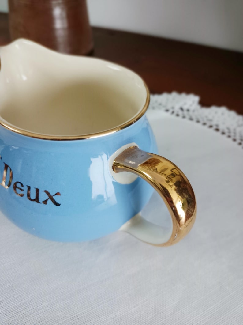 May include: A light blue creamer with a gold rim and handle. The word "Deux" is written in gold on the front of the creamer.