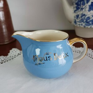 May include: A light blue creamer with a gold rim and handle. The creamer has the words "Nous Deux" written in gold lettering on the front.