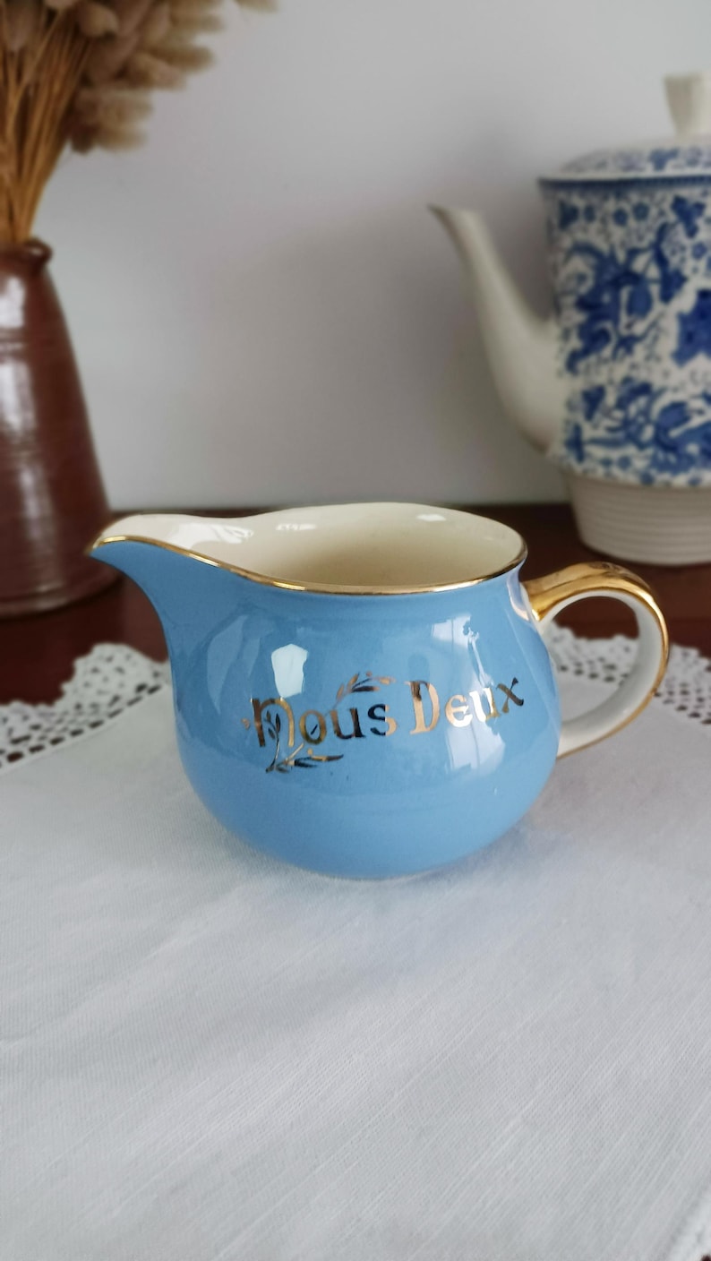 May include: A light blue creamer with a gold rim and handle. The creamer has the words "Nous Deux" written in gold lettering on the front.