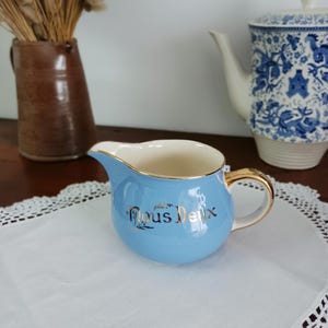 May include: A light blue creamer with a gold rim and handle. The creamer has a gold inscription that reads "Nous Deux".